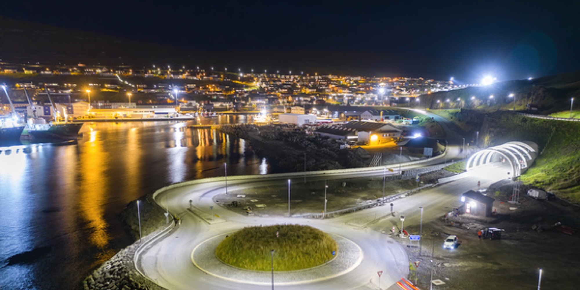 The Eysturoyartunnilin tunnel entrance and roundabout lit up at night, with the town behind